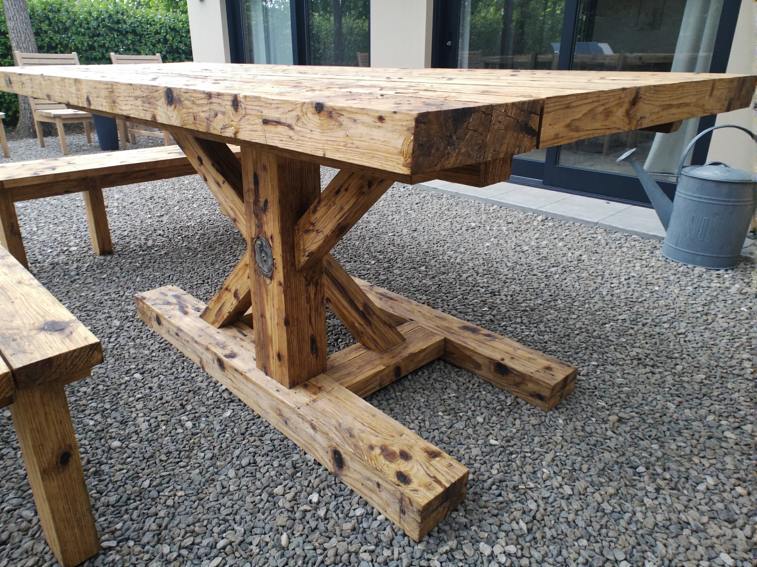 Stół ogrodowy z drewna dębowego z odzysku, otoczony zielenią i stylowymi krzesłami.